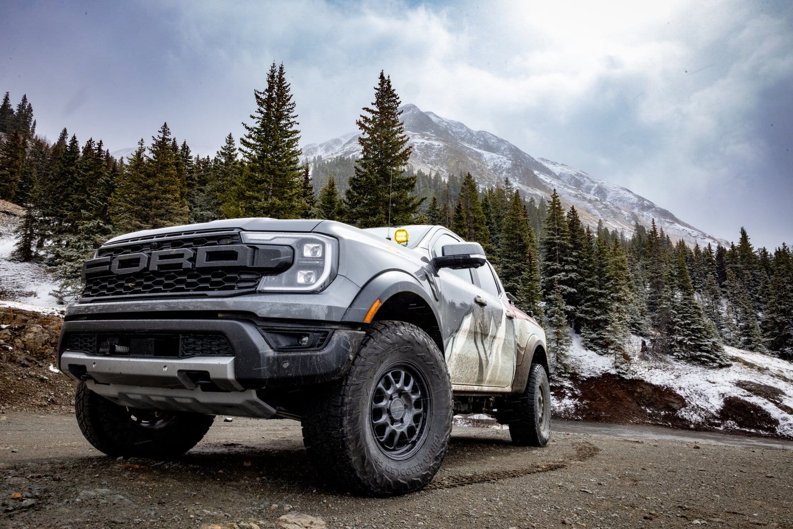 Silver Ford F-150 Raptor on mountain dirt road with dramatic snow-covered peaks and forest backdrop
