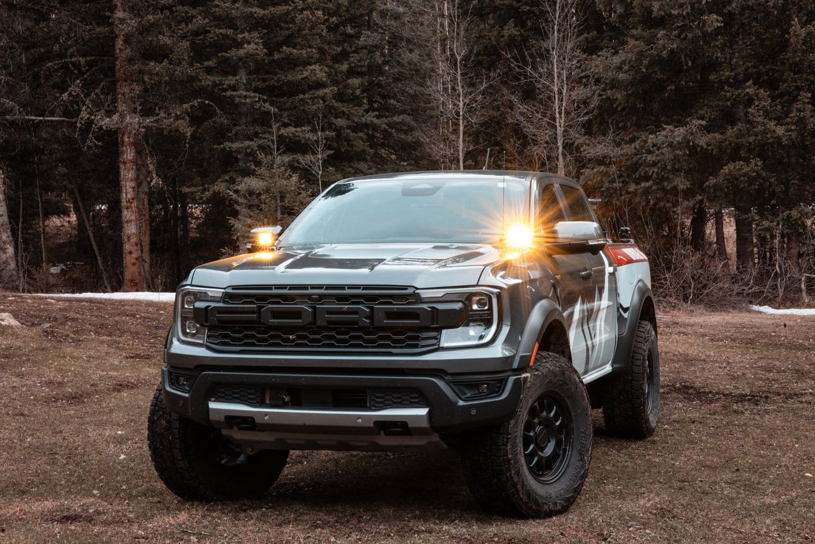 White Ford F-150 Raptor with illuminated headlights and amber lights creating lens flares in forest setting