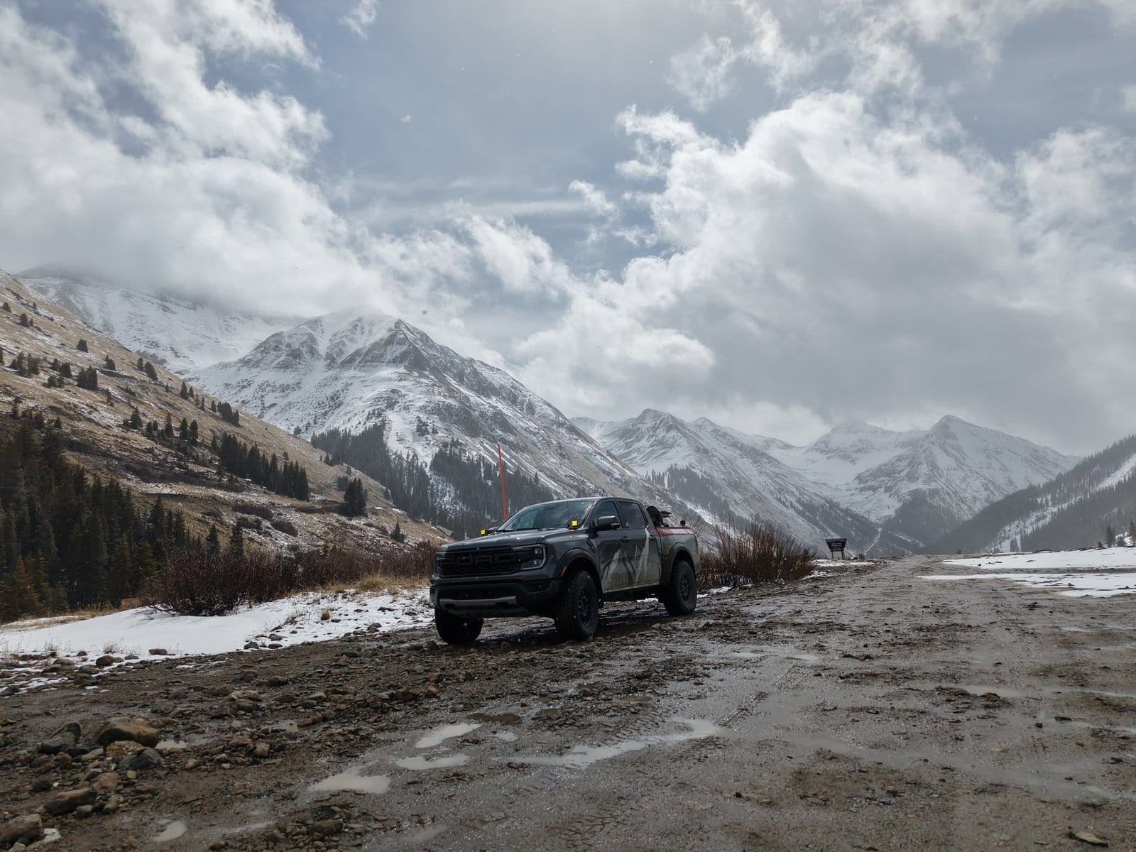 Modified Ford Raptor pickup positioned on muddy mountain trail surrounded by snow-capped alpine peaks