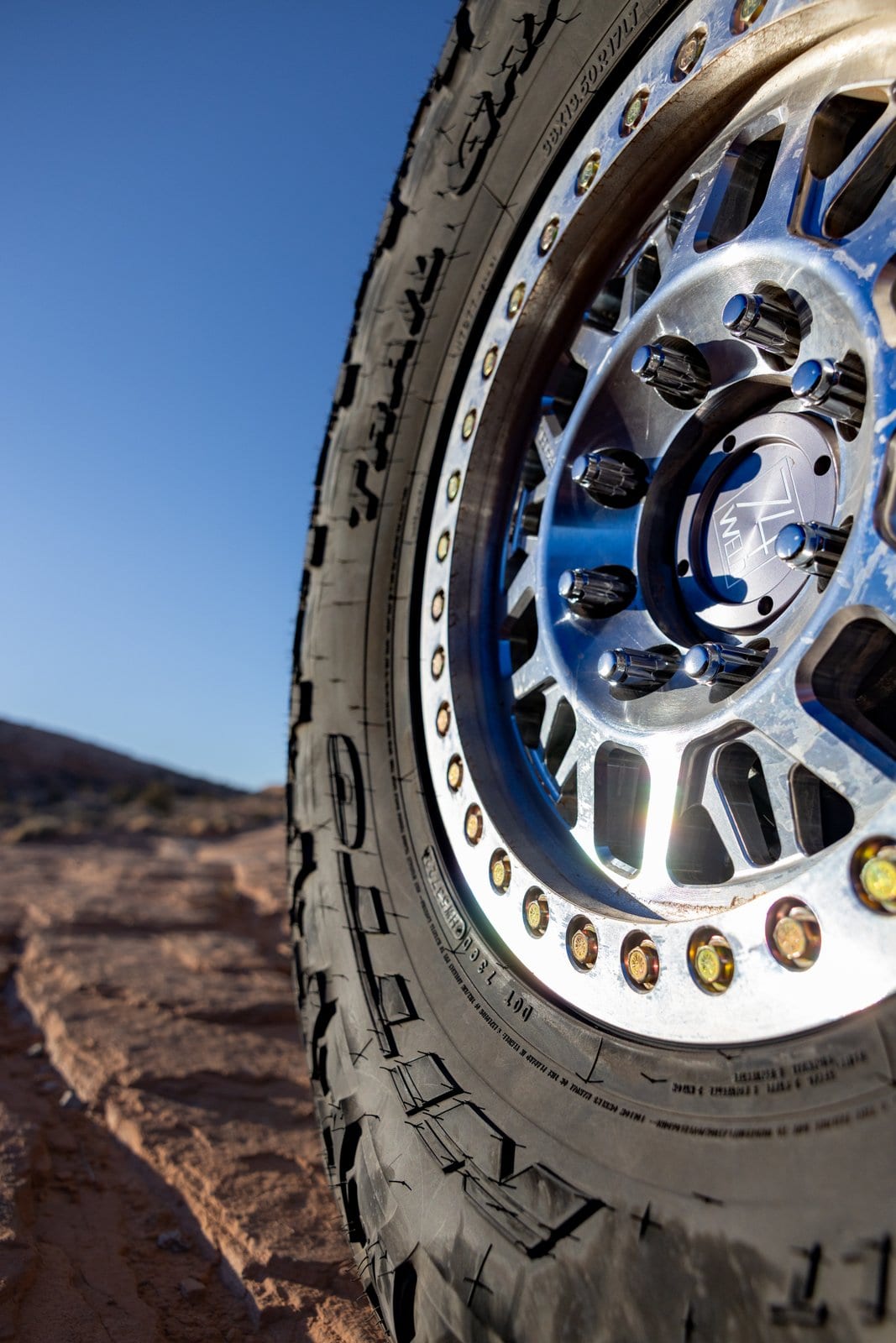 Close-up of a beadlock wheel and all-terrain tire on rocky desert ground with hills in the distance.